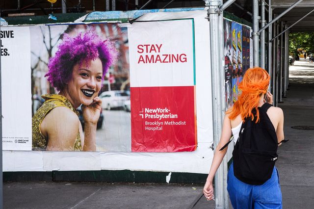 an orange-haired woman walks past a poster of a purple-haired woman